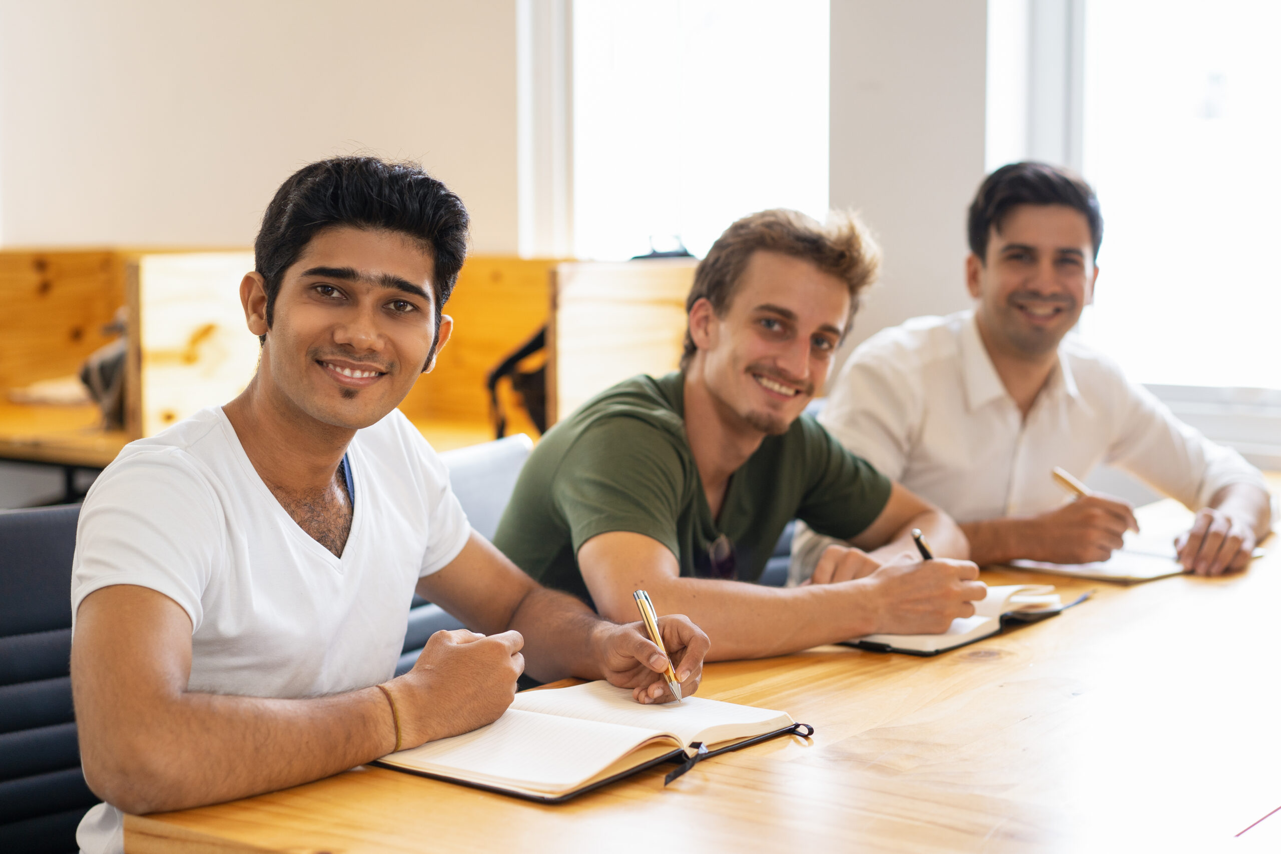 Multiethnic group of happy students posing in classroom. Three Indian and Caucasian trainees writing in notebooks and smiling at camera. Corporate education concept